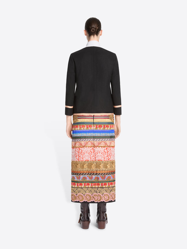 back view of a model stands against a light gray background wearing a black blazer with pink trim and a long, wide, highly patterned skirt in red, blue, gold, and beige tones, paired with dark brown boots.