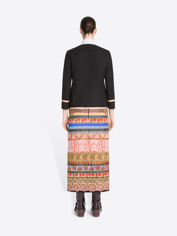 back view of a model stands against a light gray background wearing a black blazer with pink trim and a long, wide, highly patterned skirt in red, blue, gold, and beige tones, paired with dark brown boots.