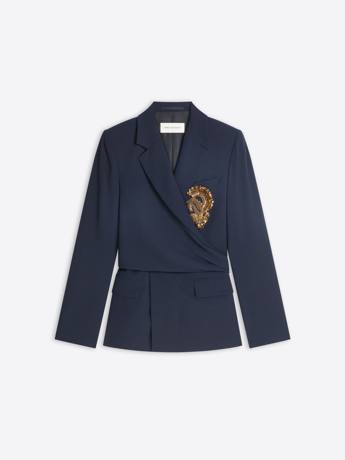 A navy blue wrap-style blazer with a structured waist and a decorative gold embroidered detail on the left chest, displayed against a light gray background.