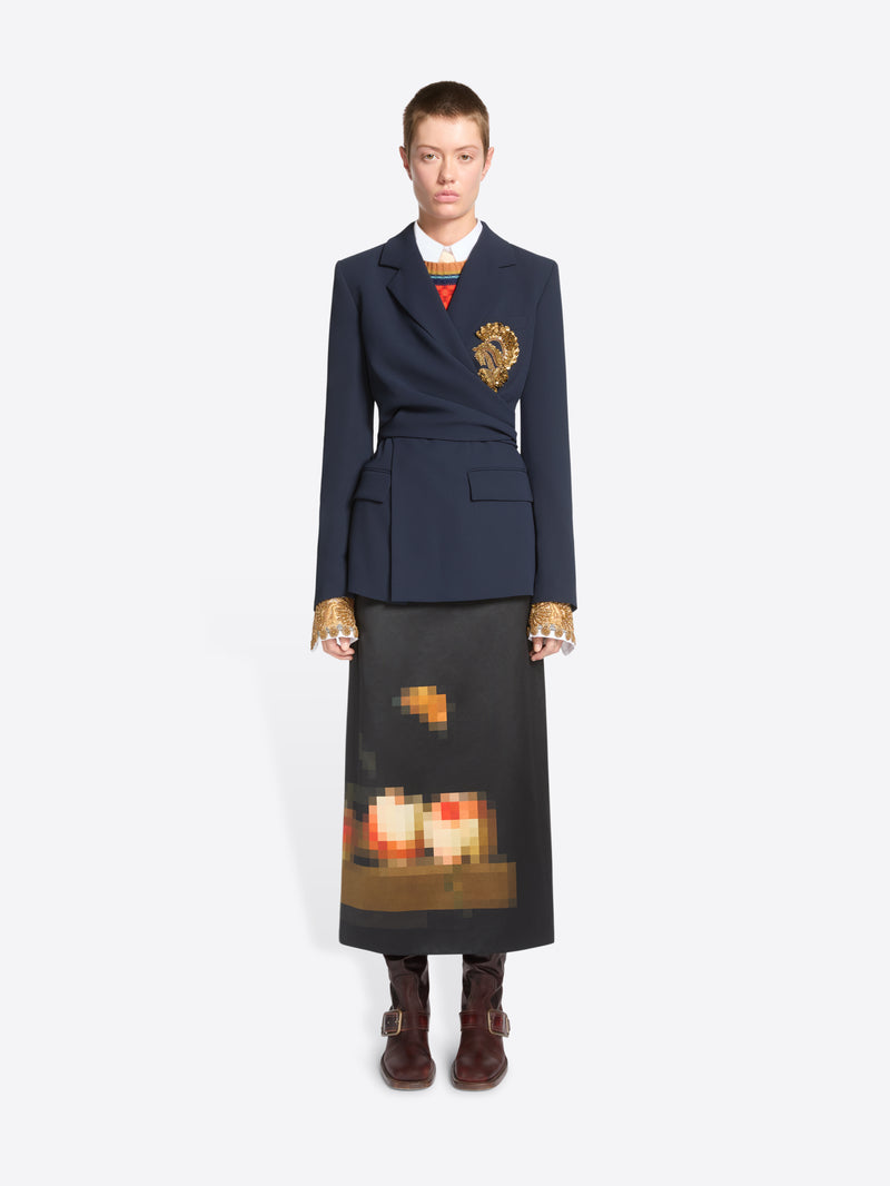 A model stands against a light gray background wearing a dark navy tailored jacket with a gold detail on the chest, a long dark skirt with a pixelated orange and yellow graphic near the hem and dark brown ankle boots.