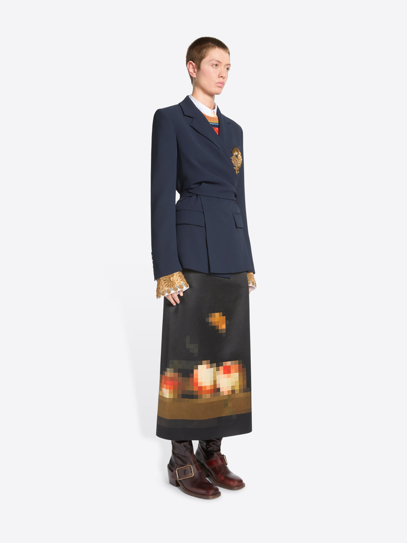 A model stands against a light gray background wearing a dark navy tailored jacket with a gold detail on the chest, a long dark skirt with a pixelated orange and yellow graphic near the hem and dark brown ankle boots.