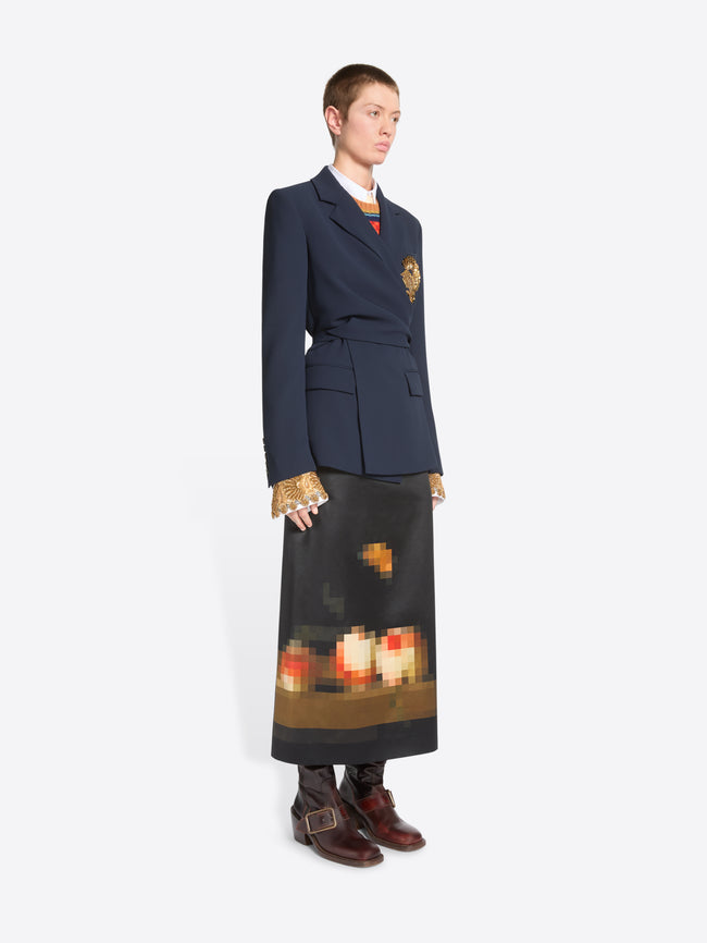 A model stands against a light gray background wearing a dark navy tailored jacket with a gold detail on the chest, a long dark skirt with a pixelated orange and yellow graphic near the hem and dark brown ankle boots.