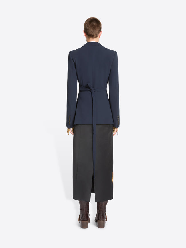 Back view of a model standing against a light gray background wearing a dark navy tailored jacket, a long dark skirt and dark brown ankle boots.