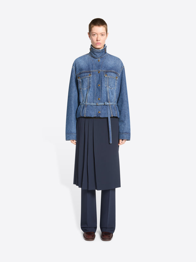 A model stands against a light gray background wearing a cropped blue denim jacket with a high mock neck, button front, and flap chest pockets, paired with a dark navy pleated skirt and matching navy trousers.