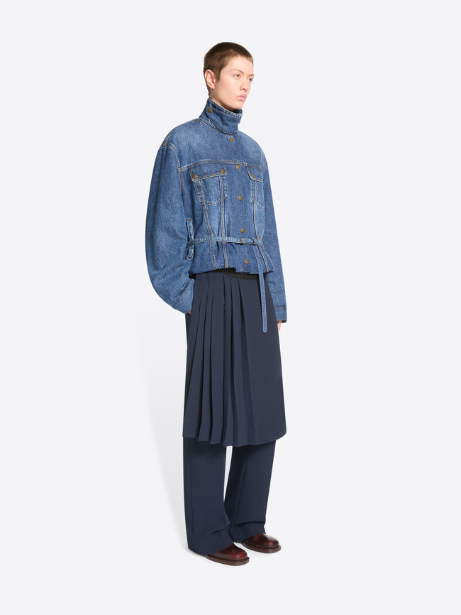 A model stands against a light gray background wearing a cropped blue denim jacket with a high mock neck, button front, and flap chest pockets, paired with a dark navy pleated skirt and matching navy trousers.