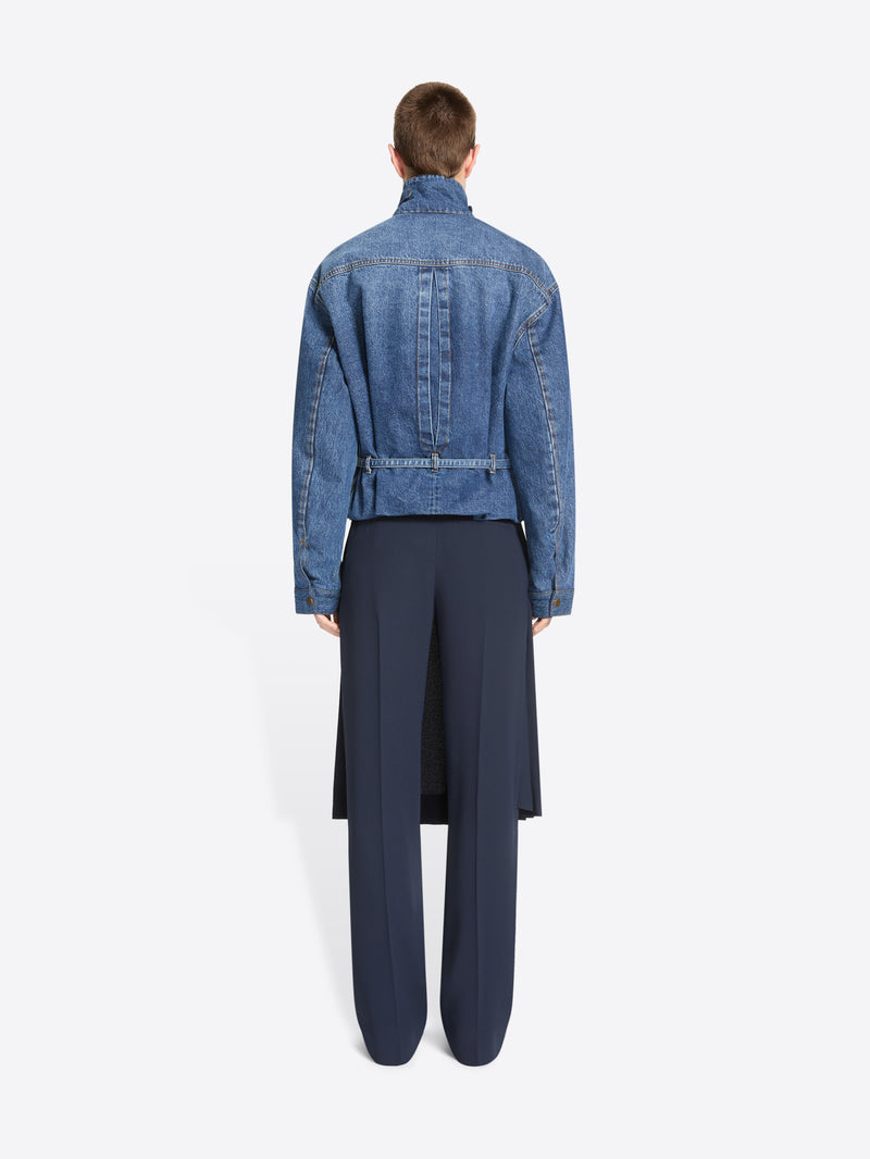 Back view of a model stands against a light gray background wearing a cropped blue denim jacket with a high mock neck, paired with a dark navy pleated skirt and matching navy trousers.