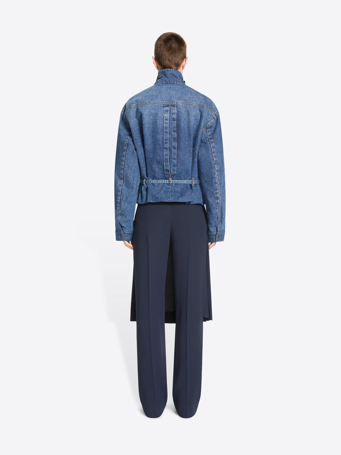 Back view of a model stands against a light gray background wearing a cropped blue denim jacket with a high mock neck, paired with a dark navy pleated skirt and matching navy trousers.