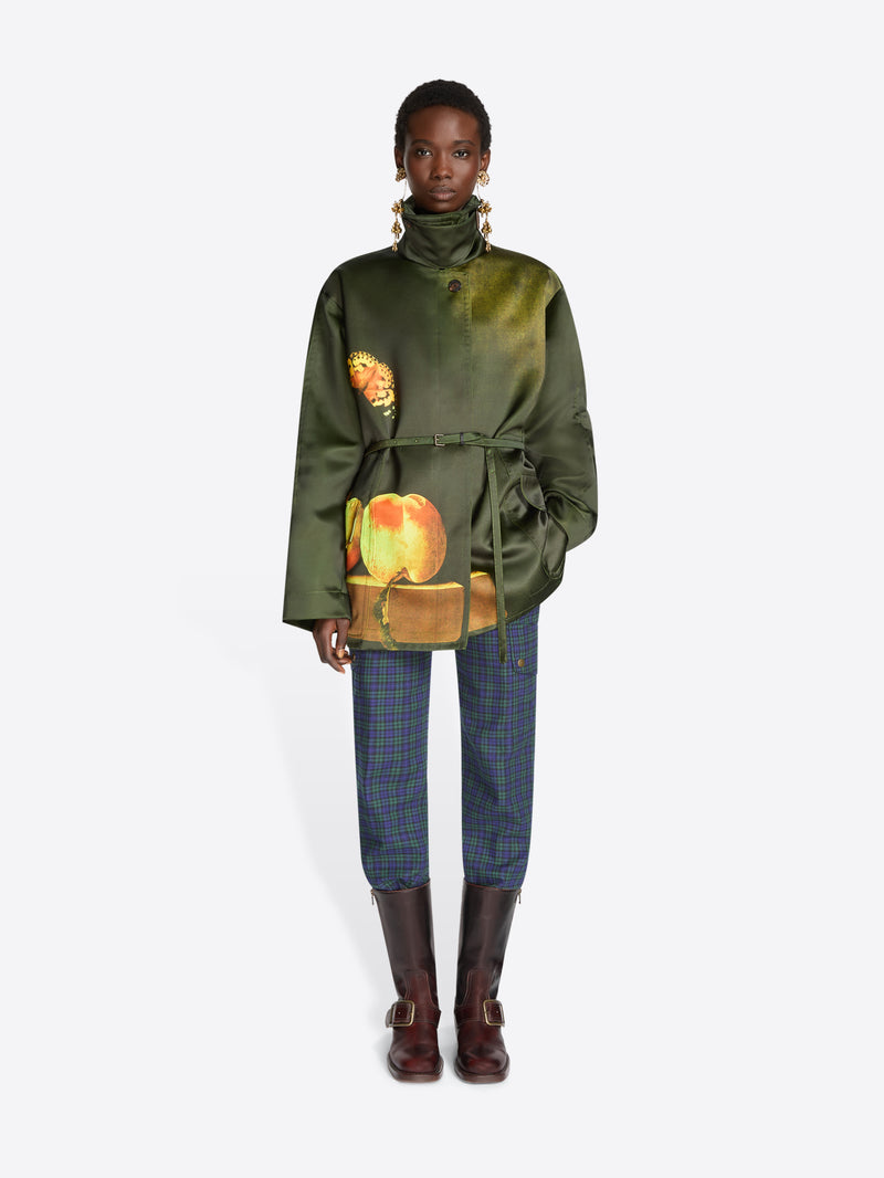 A model stands against a light gray background wearing a dark green jacket with a colorful fruit print on the front, blue plaid trousers, and dark brown boots.