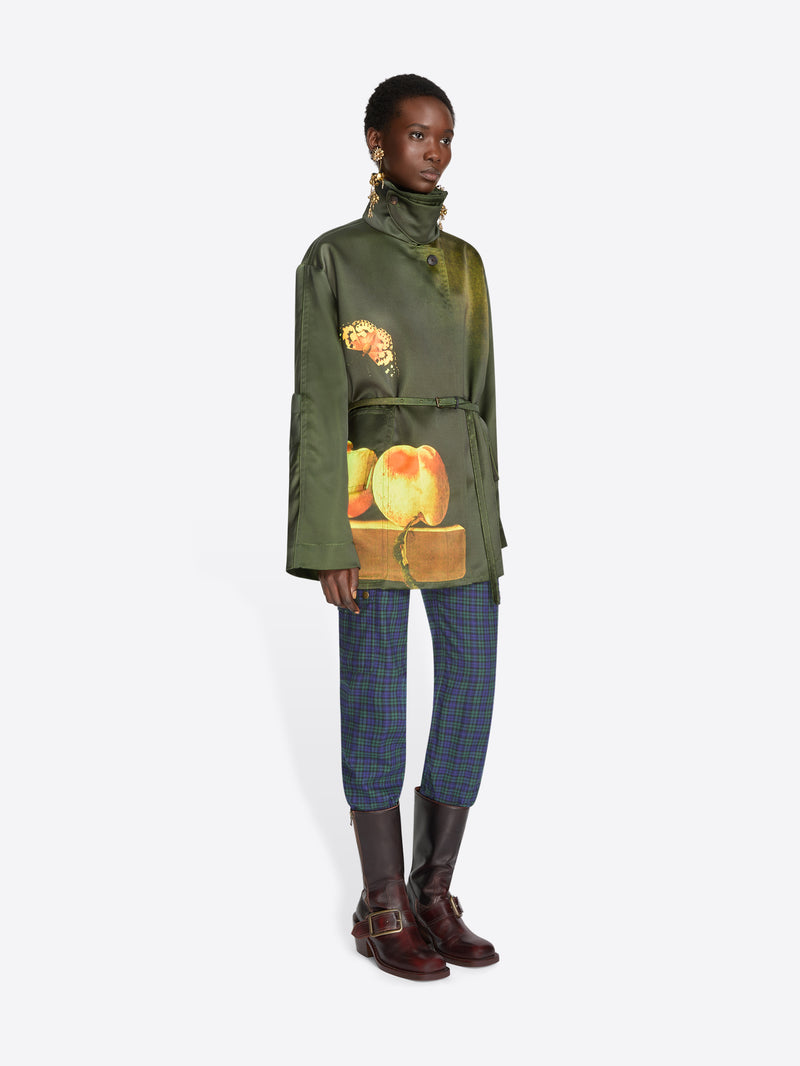 A model stands against a light gray background wearing a dark green jacket with a colorful fruit print on the front, blue plaid trousers, and dark brown boots.