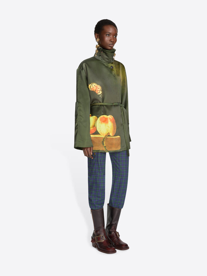 A model stands against a light gray background wearing a dark green jacket with a colorful fruit print on the front, blue plaid trousers, and dark brown boots.