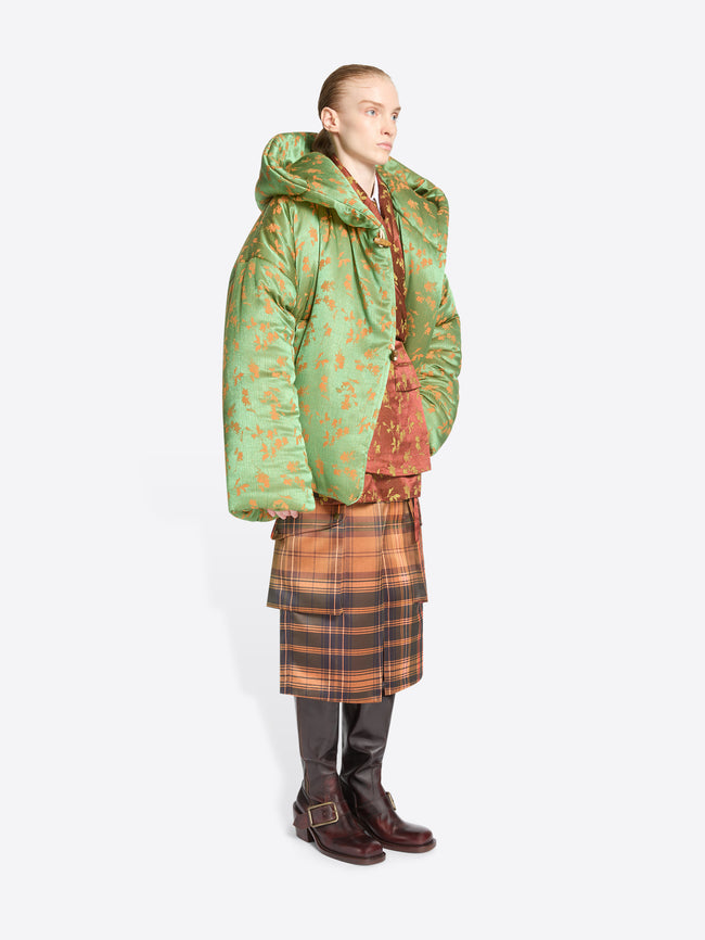 A model stands against a light gray background wearing a layered fashion outfit with a large green patterned puffer jacket over a rust-orange patterned top, a brown and orange plaid skirt, and dark brown boots.