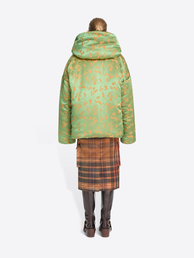 Back view of a model standing against a light gray background wearing a layered fashion outfit with a large green patterned puffer jacket over a rust-orange patterned top, a brown and orange plaid skirt, and dark brown boots.
