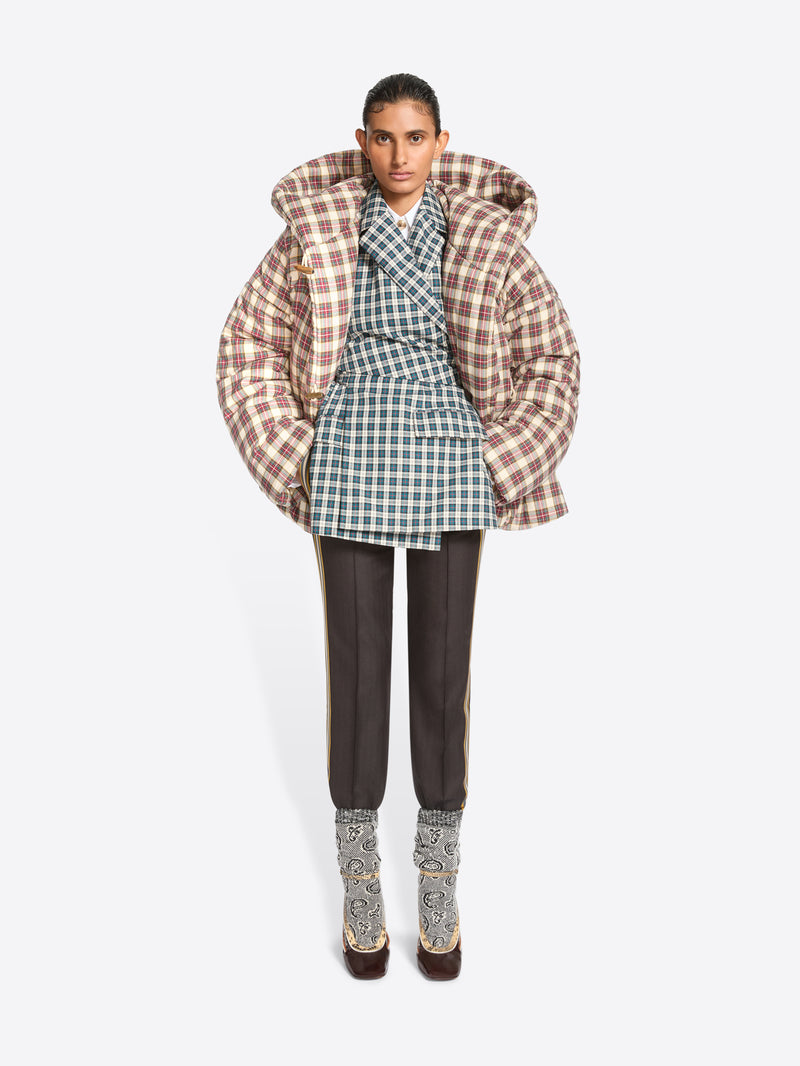 A model stands against a light gray background wearing a voluminous puffer jacket with a brown and beige plaid pattern, layered over a green and white plaid blazer top, dark trousers, and heels.