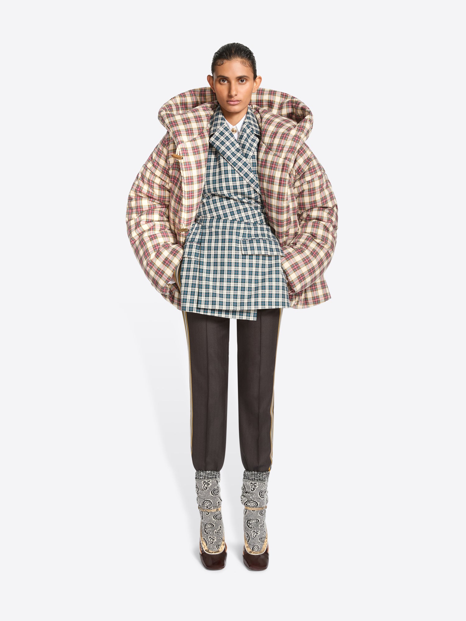 A model stands against a light gray background wearing a voluminous puffer jacket with a brown and beige plaid pattern, layered over a green and white plaid blazer top, dark trousers, and heels.