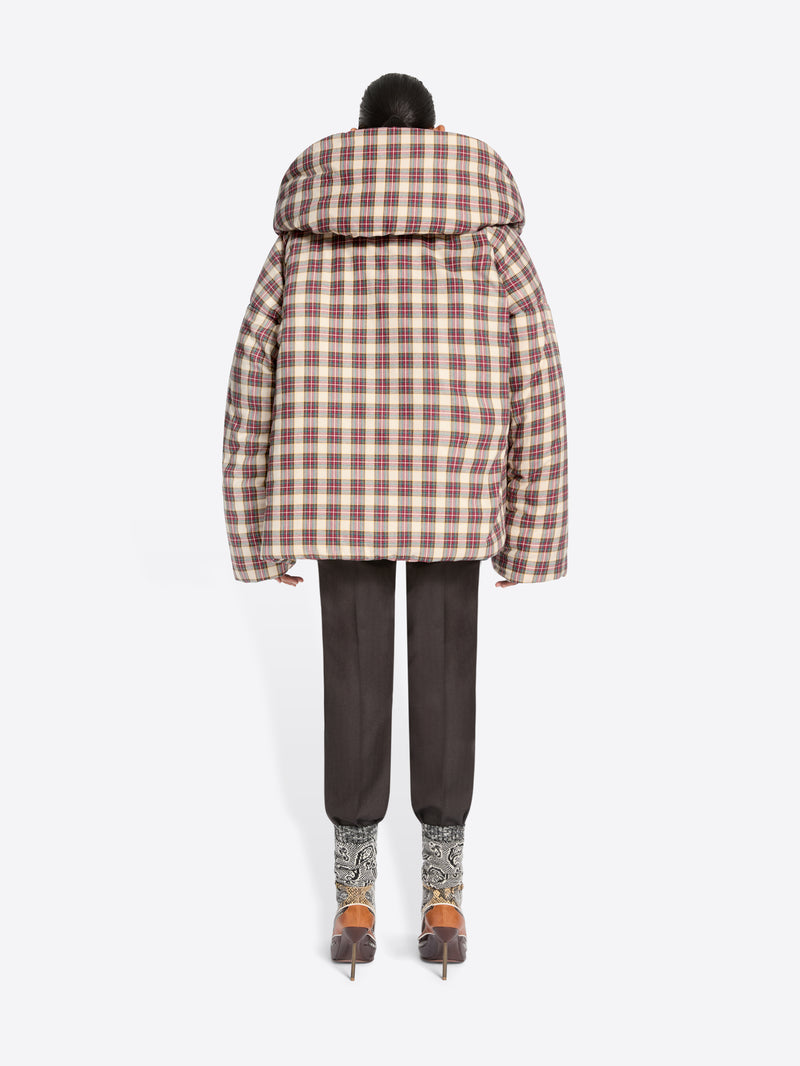 Back view of a model standing against a light gray background wearing a voluminous puffer jacket with a brown and beige plaid pattern, layered over a green and white plaid blazer top, dark trousers, and heels.