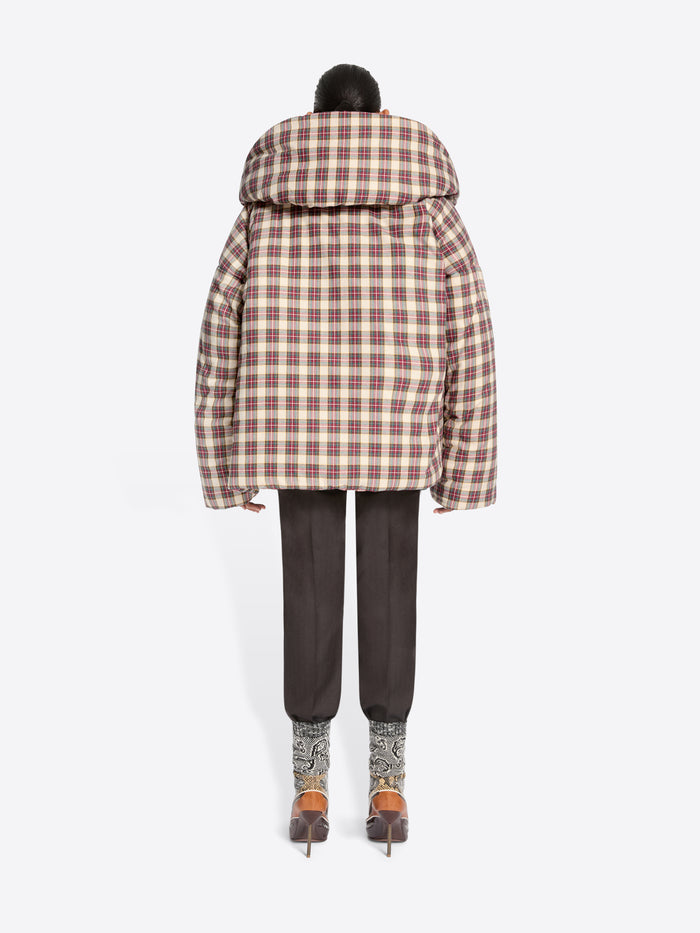 Back view of a model standing against a light gray background wearing a voluminous puffer jacket with a brown and beige plaid pattern, layered over a green and white plaid blazer top, dark trousers, and heels.