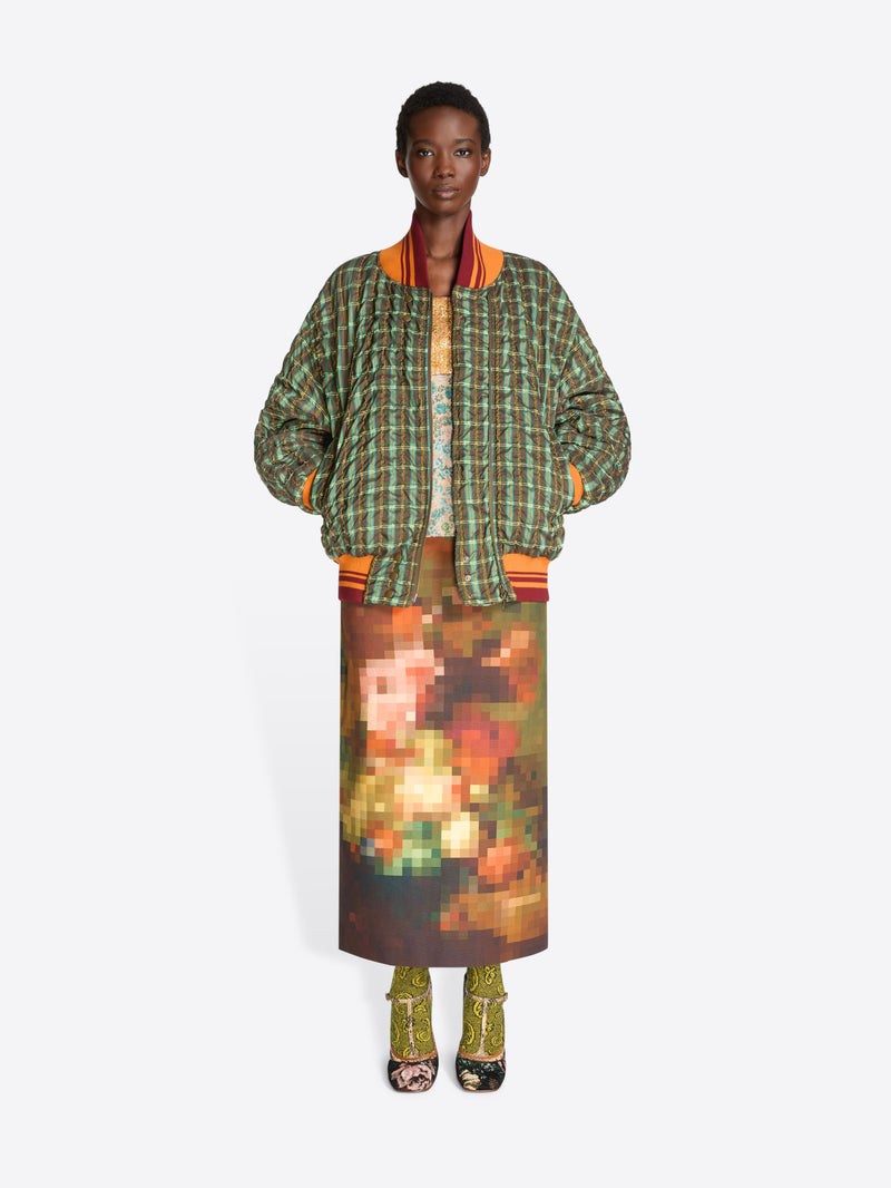 A model stands against a light gray background wearing a green and orange plaid bomber jacket with a top underneath, a brown and orange pixelated-patterned skirt and heels.