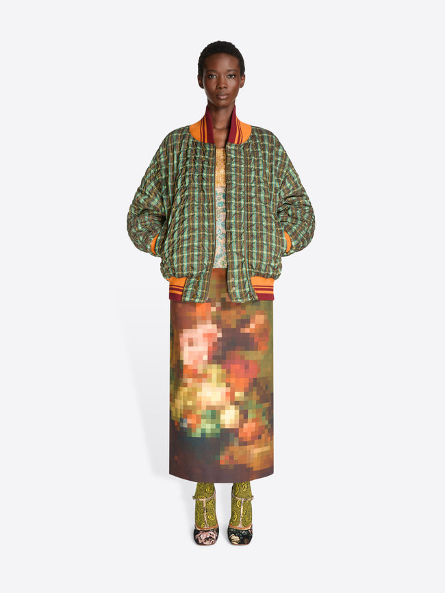 A model stands against a light gray background wearing a green and orange plaid bomber jacket with a top underneath, a brown and orange pixelated-patterned skirt and heels.