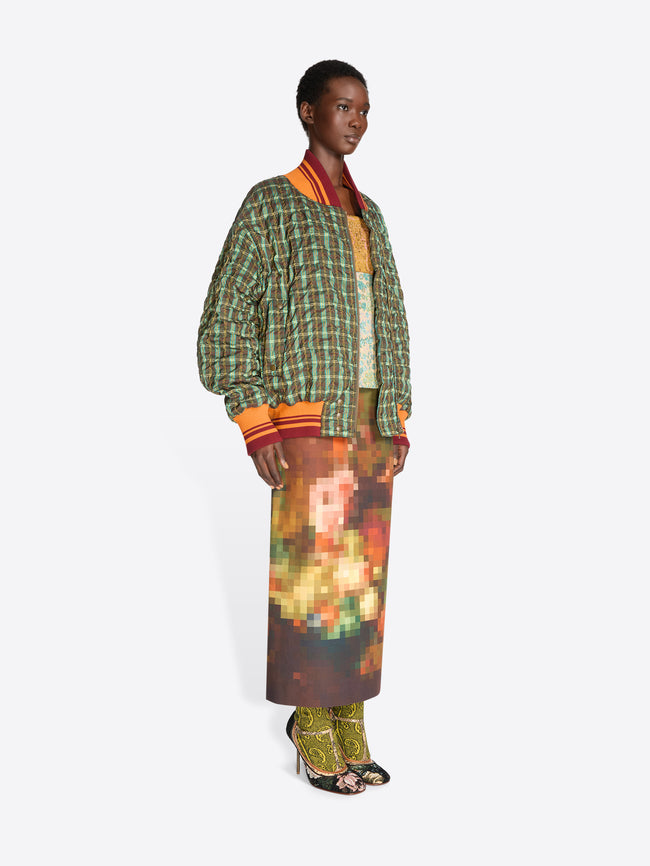 A model stands against a light gray background wearing a green and orange plaid bomber jacket with a top underneath, a brown and orange pixelated-patterned skirt and heels.
