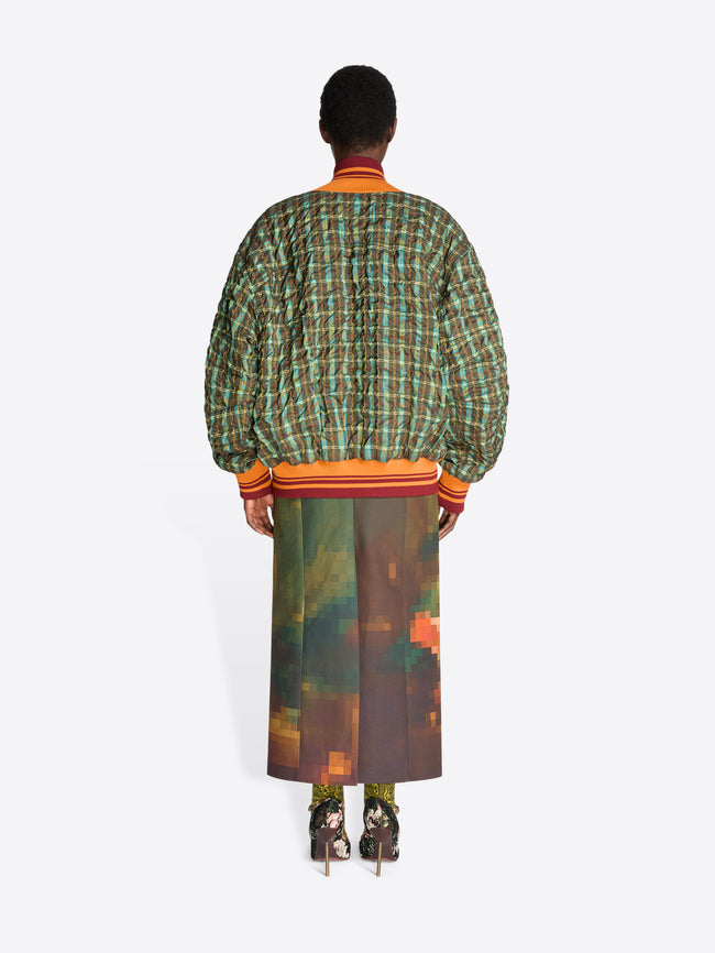 Back view of a model standing against a light gray background wearing a green and orange plaid bomber jacket with a top underneath, a brown and orange pixelated-patterned skirt and heels.