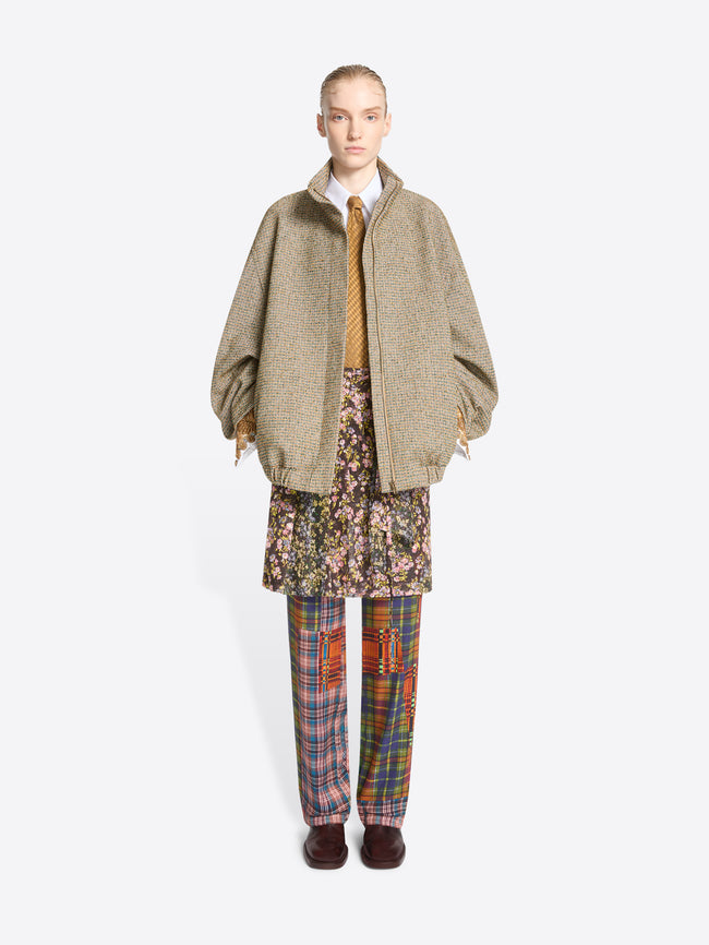 A model stands against a light gray background wearing a beige oversized zip-front jacket over a white collared shirt and mustard tie, paired with a floral mini skirt and multicolor plaid trousers.