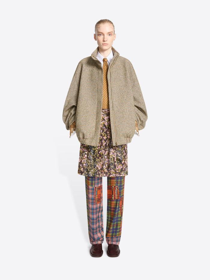 A model stands against a light gray background wearing a beige oversized zip-front jacket over a white collared shirt and mustard tie, paired with a floral mini skirt and multicolor plaid trousers.