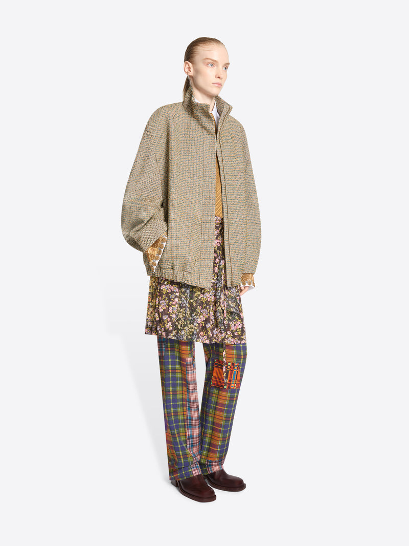 A model stands against a light gray background wearing a beige oversized zip-front jacket over a white collared shirt and mustard tie, paired with a floral mini skirt and multicolor plaid trousers.