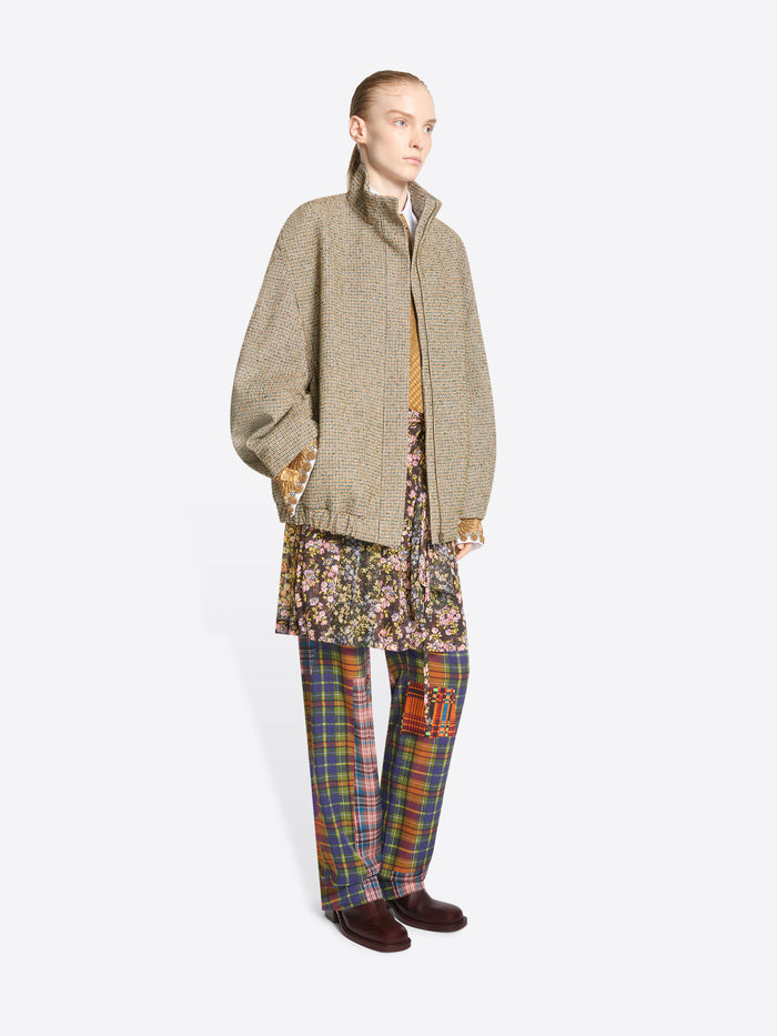A model stands against a light gray background wearing a beige oversized zip-front jacket over a white collared shirt and mustard tie, paired with a floral mini skirt and multicolor plaid trousers.