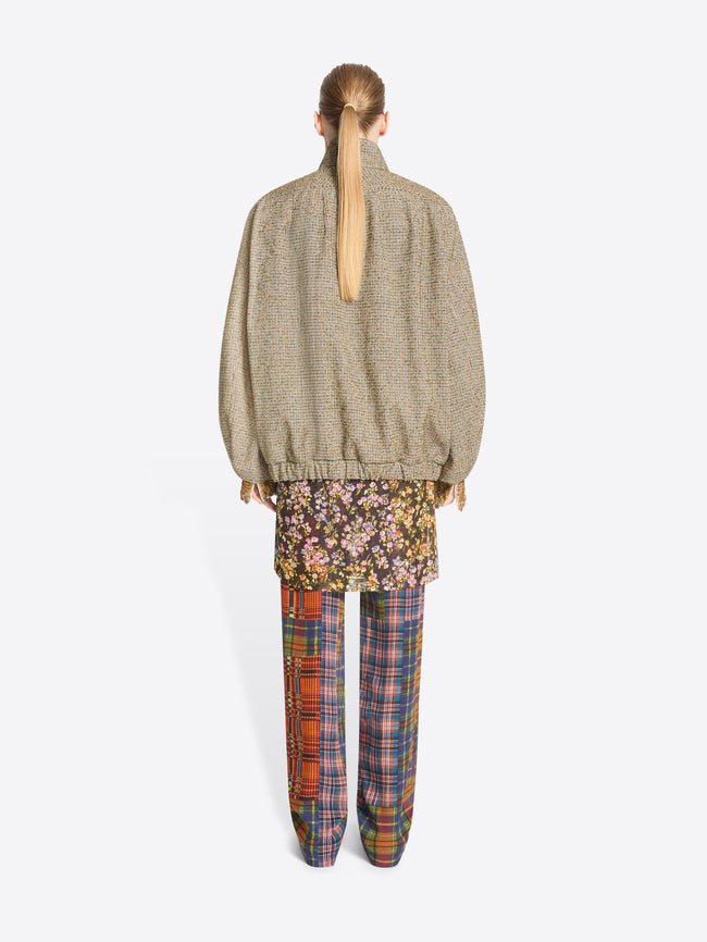 Back view of a model standing against a light gray background wearing a beige oversized zip-front jacket paired with a floral mini skirt and multicolor plaid trousers.
