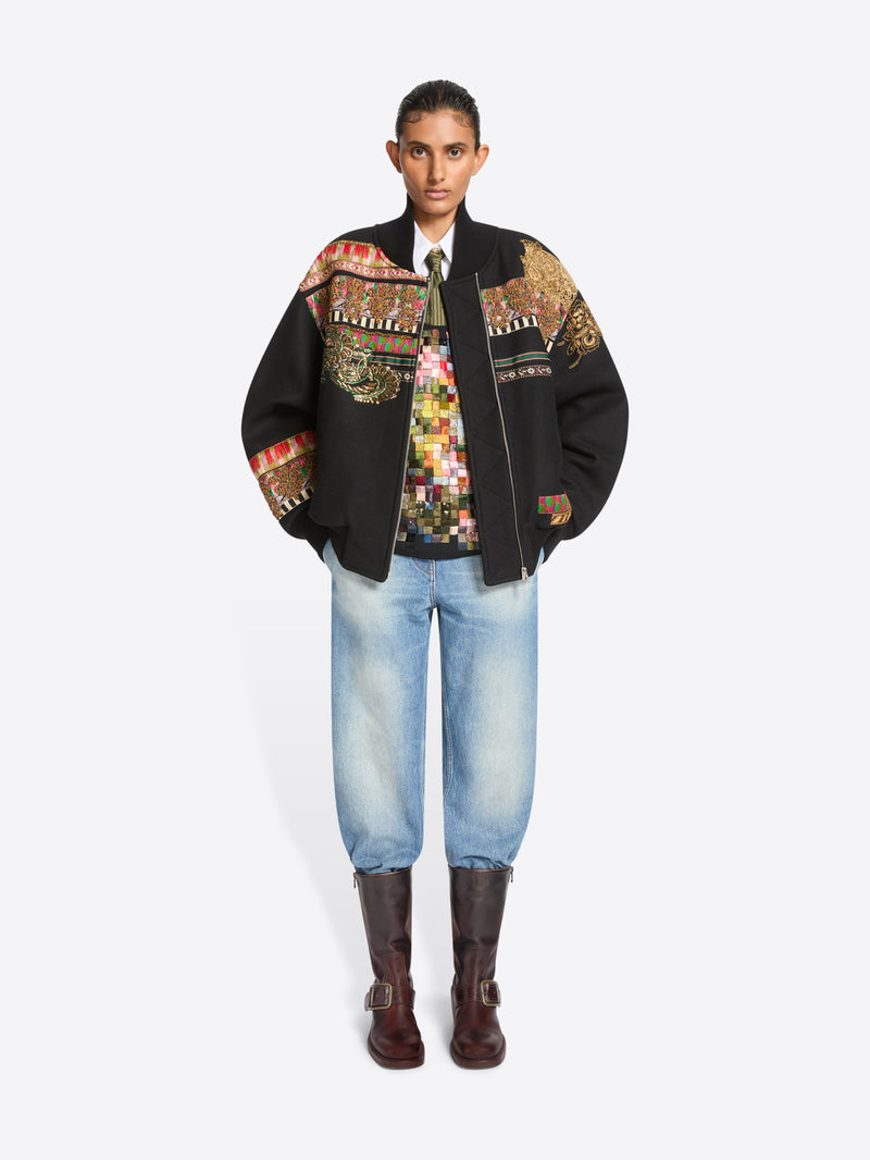 A model stands against a light gray background wearing a black bomber jacket with intricate multicolor geometric embroidery across the shoulders and sleeves, a patterned shirt, light blue jeans, and dark brown boots.