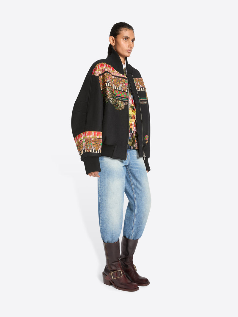 A model stands against a light gray background wearing a black bomber jacket with intricate multicolor geometric embroidery across the shoulders and sleeves, a patterned shirt, light blue jeans, and dark brown boots.