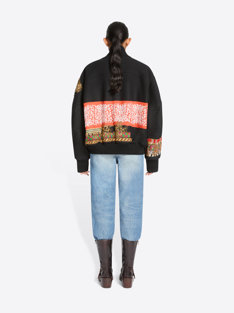 Back view of a model standing against a light gray background wearing a black bomber jacket with intricate multicolor geometric embroidery across the shoulders and sleeves, a patterned shirt, light blue jeans, and dark brown boots.