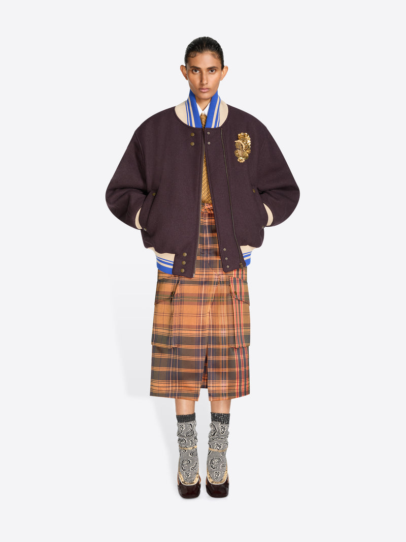A model stands against a light gray background wearing a dark plum varsity-style jacket with a gold embroidered detail, layered over a striped blue and white shirt, a brown and orange plaid midi skirt, patterned socks and dark shoes.