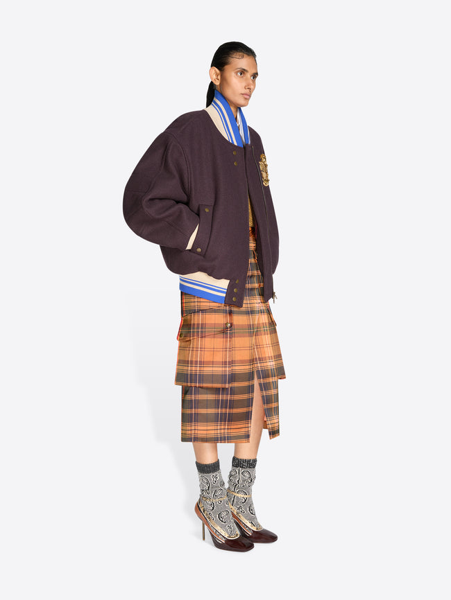 A model stands against a light gray background wearing a dark plum varsity-style jacket with a gold embroidered detail, layered over a striped blue and white shirt, a brown and orange plaid midi skirt, patterned socks and dark shoes.