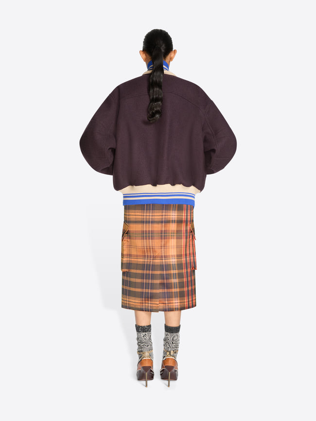 Back view of a model standing against a light gray background wearing a dark plum varsity-style jacket, a brown and orange plaid midi skirt, patterned socks and heels.