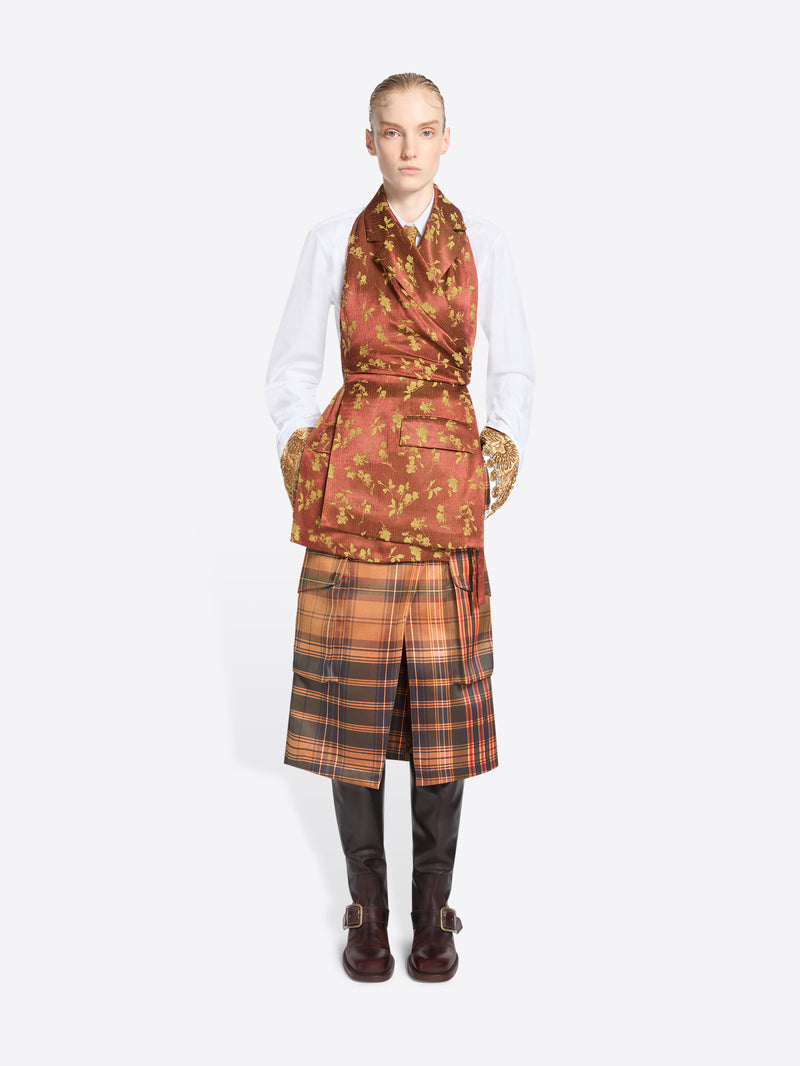 A model stands against a light gray background wearing a white shirt, a rust-orange patterned top with, a brown plaid skirt, and dark brown boots.