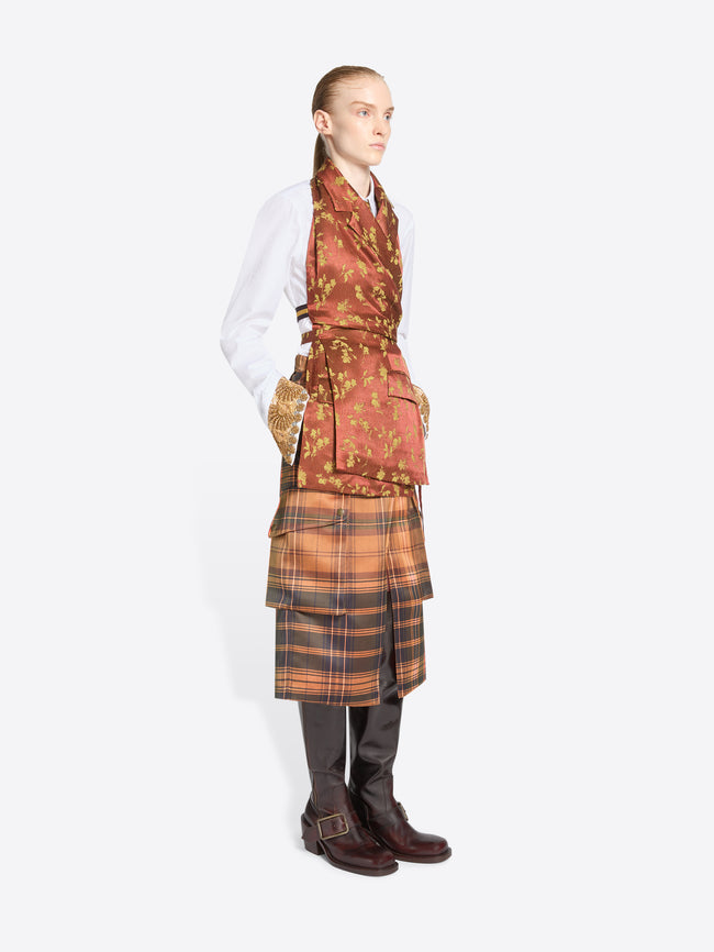 A model stands against a light gray background wearing a white shirt, a rust-orange patterned top with, a brown plaid skirt, and dark brown boots.