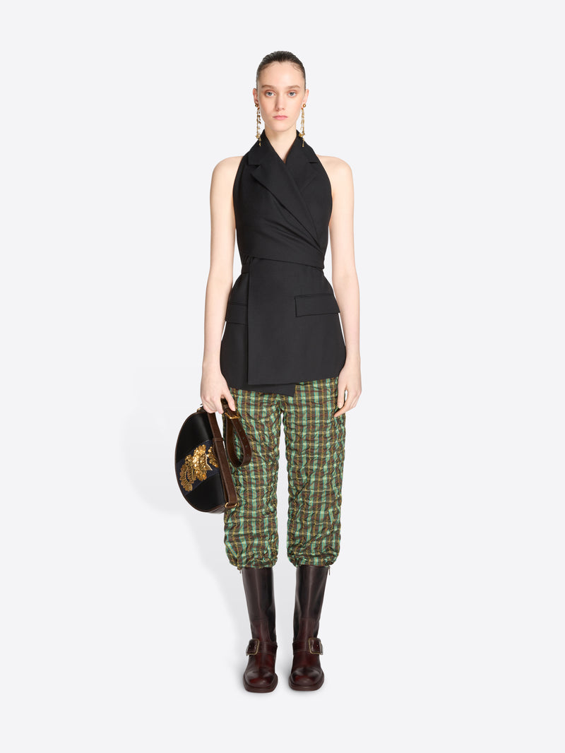 A model stands against a light gray background wearing a sleeveless black wrap-style top, green and beige plaid pants, dark brown knee-high boots, and holding a black bag with a gold detail.