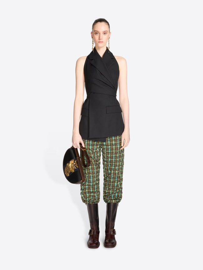 A model stands against a light gray background wearing a sleeveless black wrap-style top, green and beige plaid pants, dark brown knee-high boots, and holding a black bag with a gold detail.