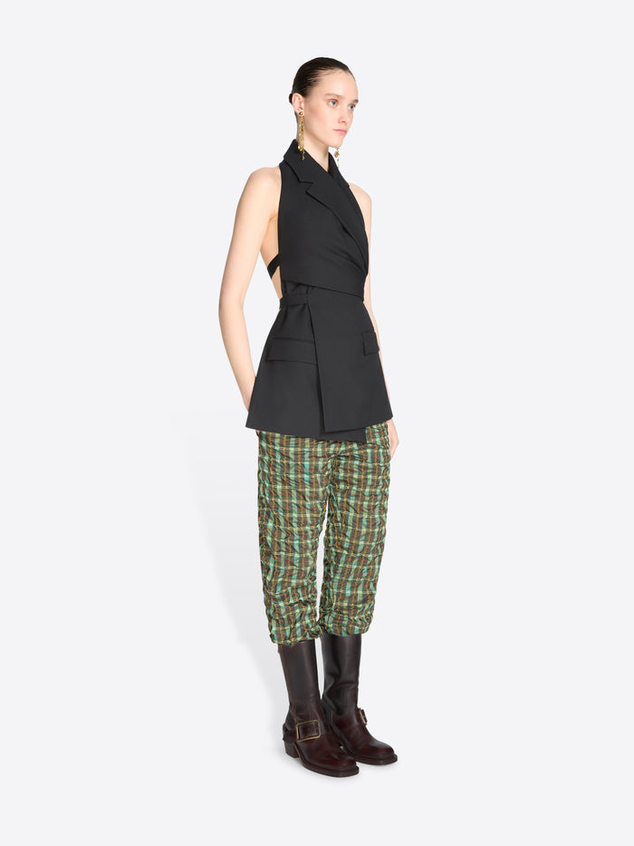 A model stands against a light gray background wearing a sleeveless black wrap-style top, green and beige plaid pants, dark brown knee-high boots.