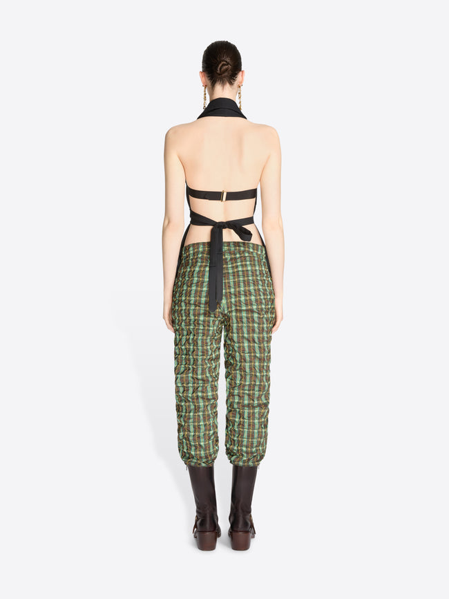Back view of a model standing against a light gray background wearing a sleeveless black wrap-style top, green and beige plaid pants, dark brown knee-high boots.