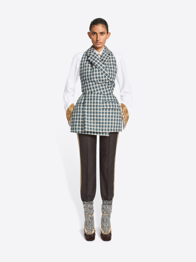 A model stands against a light gray background wearing a white long-sleeve shirt, a green and white checkered wrap-style top, dark brown pants, and high-heels.