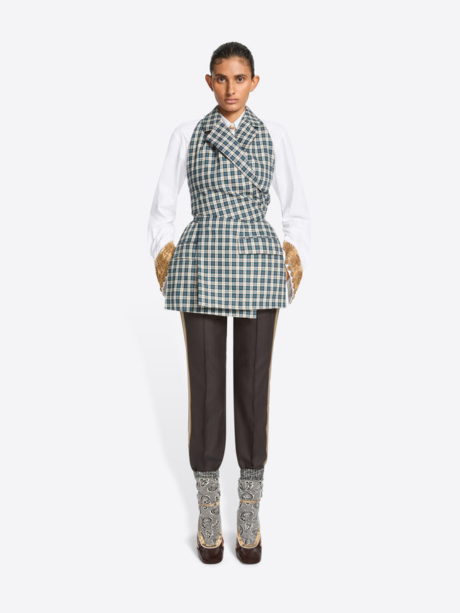 A model stands against a light gray background wearing a white long-sleeve shirt, a green and white checkered wrap-style top, dark brown pants, and high-heels.