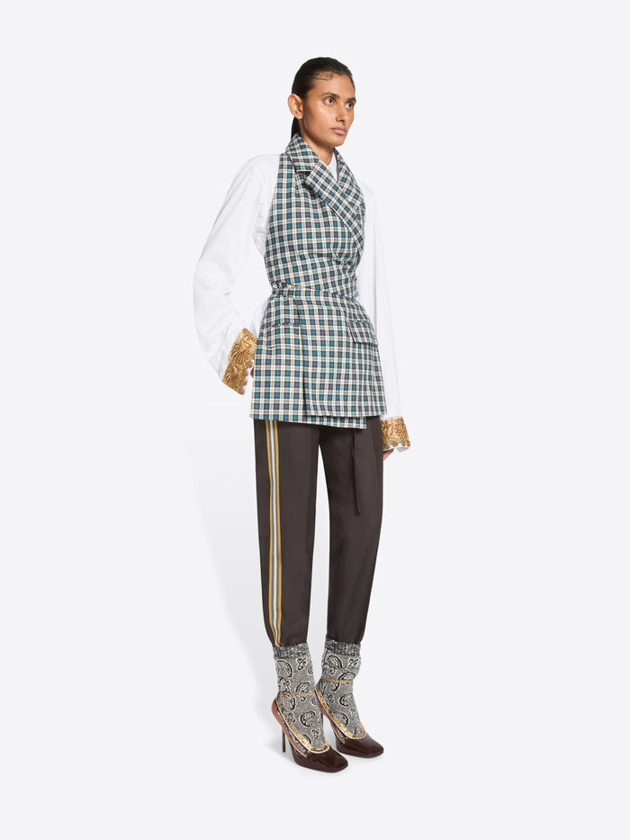 A model stands against a light gray background wearing a white long-sleeve shirt, a green and white checkered wrap-style top, dark brown pants, and high-heels.