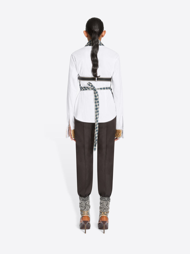 Back view of a model standing against a light gray background wearing a white long-sleeve shirt, a green and white checkered wrap-style top, dark brown pants, and high-heels.