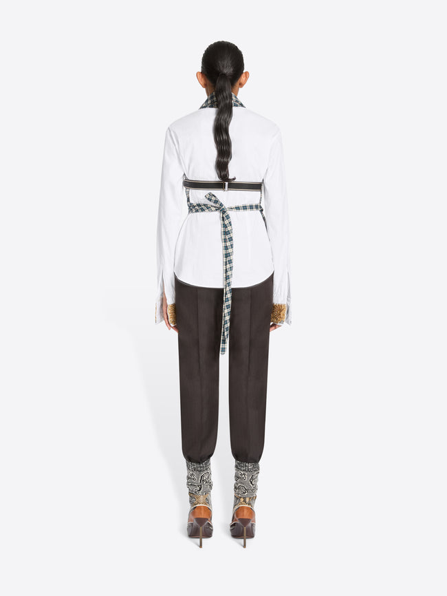 Back view of a model standing against a light gray background wearing a white long-sleeve shirt, a green and white checkered wrap-style top, dark brown pants, and high-heels.