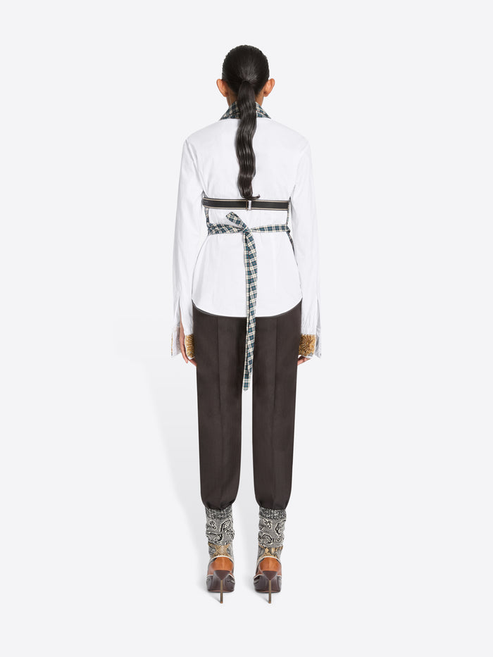 Back view of a model standing against a light gray background wearing a white long-sleeve shirt, a green and white checkered wrap-style top, dark brown pants, and high-heels.
