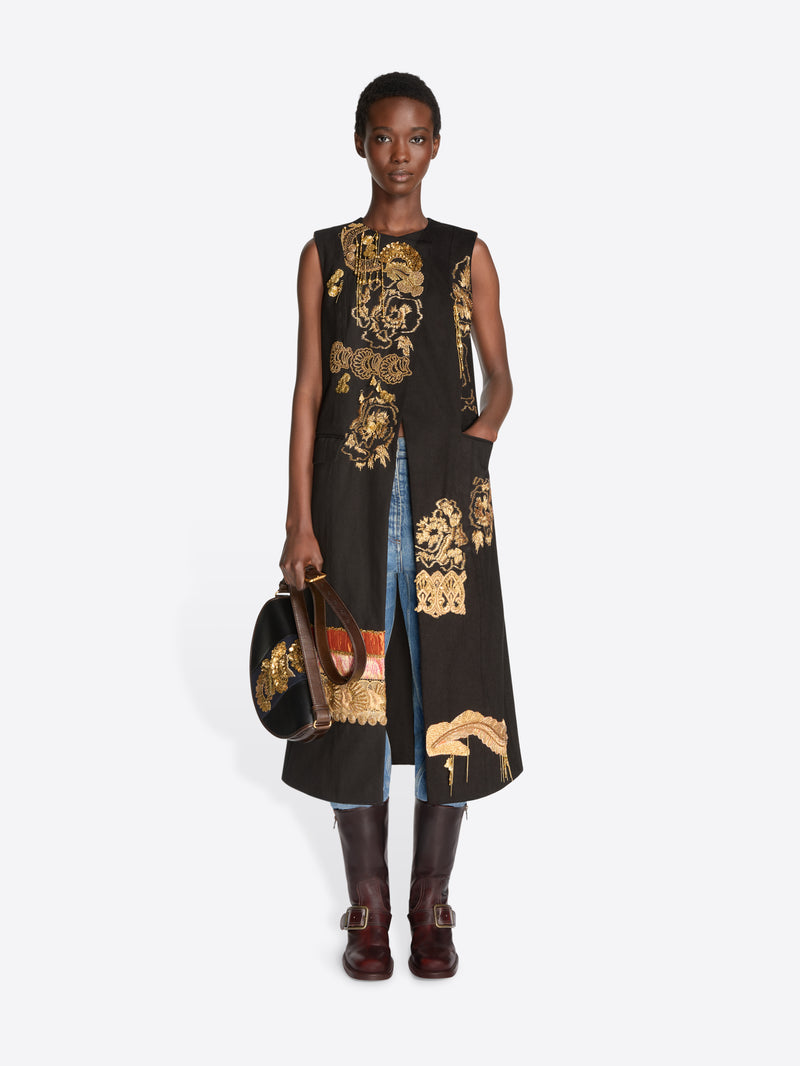A model stands against a light gray background wearing a sleeveless black coat with ornate gold baroque-style patterns, layered over light blue jeans, holding a matching black and gold patterned bag and wearing dark brown leather boots.