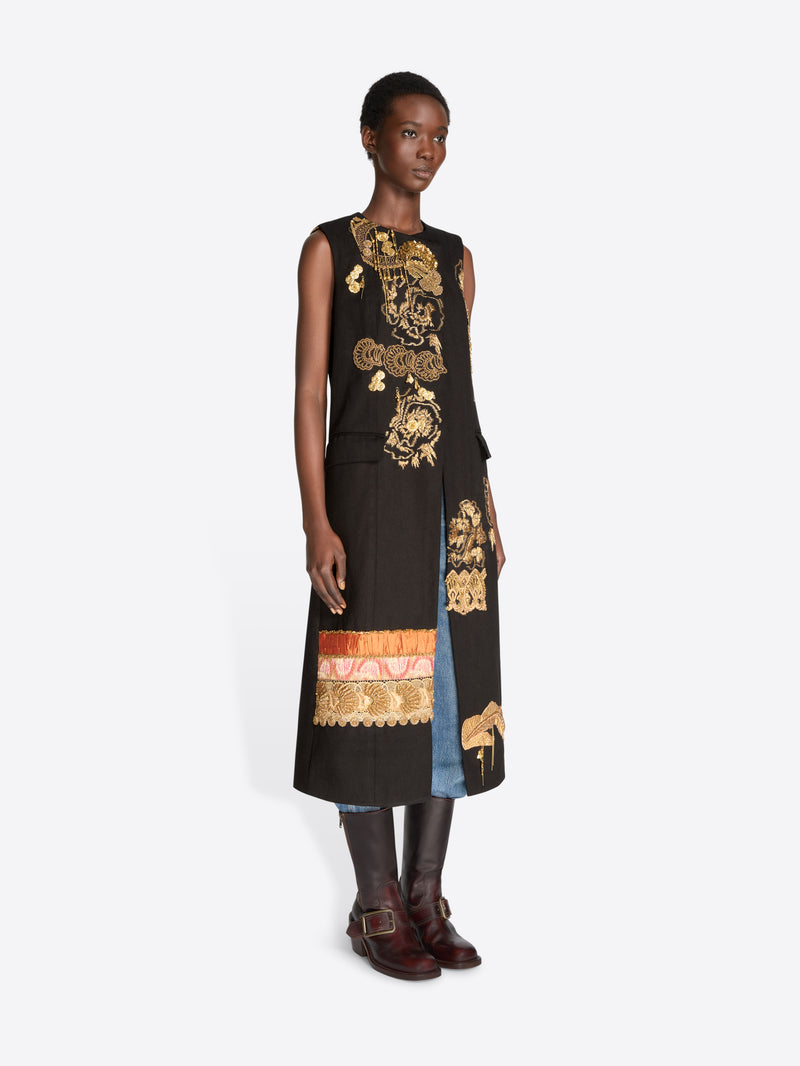 A model stands against a light gray background wearing a sleeveless black coat with ornate gold baroque-style patterns, layered over light blue jeans, holding a matching black and gold patterned bag and wearing dark brown leather boots.