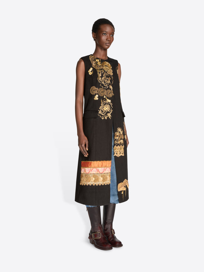 A model stands against a light gray background wearing a sleeveless black coat with ornate gold baroque-style patterns, layered over light blue jeans, holding a matching black and gold patterned bag and wearing dark brown leather boots.